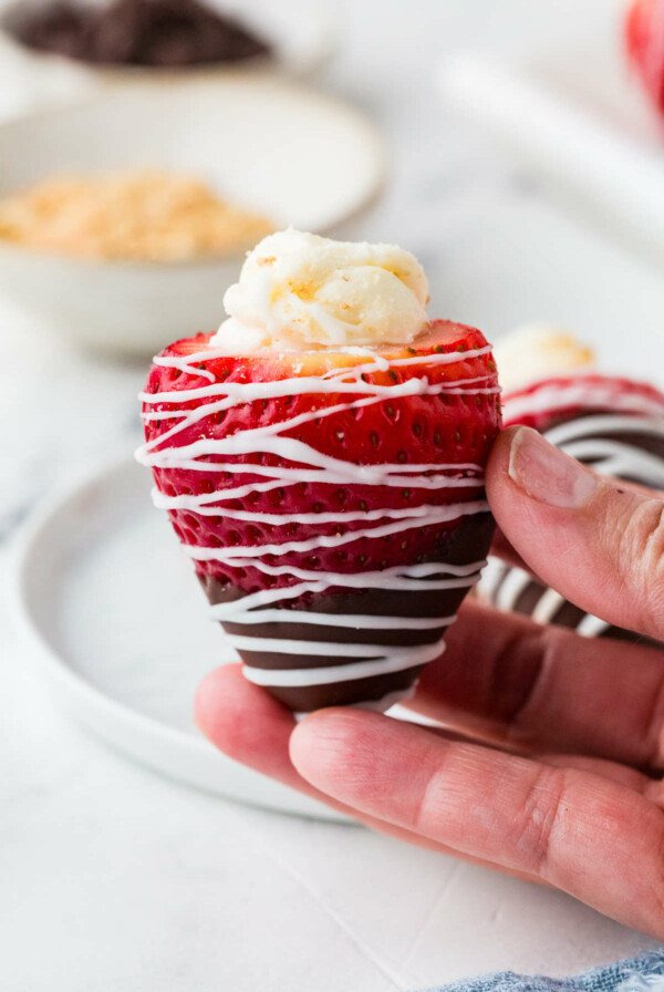 A hand holding a strawberry stuffed with cheesecake filling and dipped in chocolate.